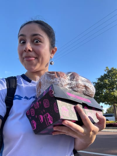 A person, likely female, is holding a box of baked goods wrapped in plastic in an outdoor setting. The box is black with pink dessert-themed designs. The person has dark hair pulled back, is wearing a white shirt with blue details, and has dice-shaped earrings. A backpack strap is visible. Background features include blue sky, power lines, a tree, and a glimpse of a building or vehicle. There is no direct indication of Bassendean, Australia in the picture.