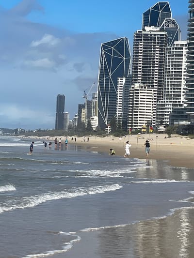 This is a photograph taken at Surfers Paradise, Australia, depicting a beach scene with a backdrop of modern high-rise buildings. The foreground is dominated by the ocean with gentle waves lapping at the sandy shore. Several people are visible on the beach and in the shallow water. In the distance, a cluster of tall buildings, characteristic of the Surfers Paradise skyline, rises against a partly cloudy blue sky. Construction cranes are also visible amongst the buildings, suggesting ongoing development. On the beach, some individuals appear to be walking or standing, while others are interacting in smaller groups. The overall impression is of a typical day at a popular urban beach.