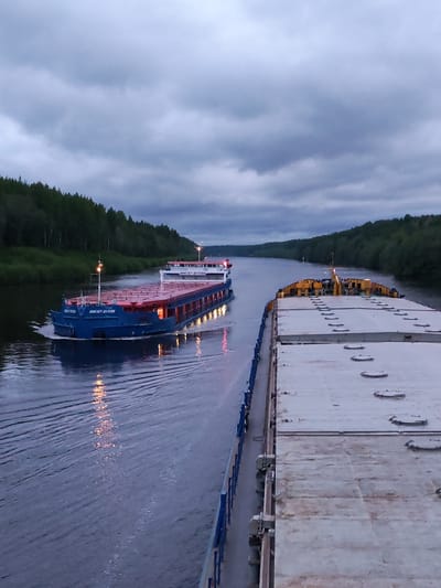 The image shows two barges on a canal. The barge in the foreground is primarily gray with some rust, while the barge in the background is blue and red, with lights reflecting on the water. Both vessels are heading in opposite directions. The scene is framed by trees lining the canal banks and a cloudy, overcast sky. The canal location is in the area of Aleksandrovskoe, Russia. There are no people in view.