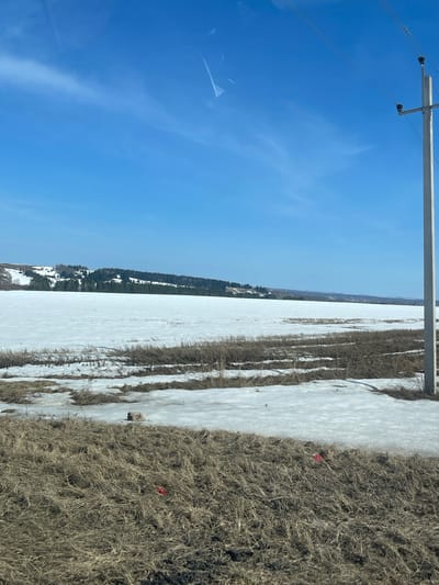 The image depicts a landscape in Kueda, Russia, during a period of snow cover. A power line infrastructure is visible in the upper left. The ground transitions from snow-covered fields to areas of dry, brown vegetation. In the distance, a line of trees is present. A red object is discernible amidst the dry vegetation. The sky is blue with wisps of clouds.