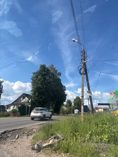 The image depicts a street scene in Medyn, Russia, on a clear day with a blue sky and scattered clouds. A white SUV is driving on a paved road that shows signs of disrepair, with a pothole and debris along its edge. To the left of the road, a light-colored house with a gable roof is visible, surrounded by trees and a fence. In the foreground on the right, tall green grass and vegetation grow, with a concrete utility pole and overhead power lines dominating the center of the frame. A streetlamp is attached to the pole. In the background, several other vehicles, including a white van and a dark truck, are seen on the road. A building with green and white signage, possibly a store, is situated further down the street. A red circular sign with a white number "5" is affixed to the building. The overall environment suggests a rural or semi-urban setting with a mix of residential and commercial elements.