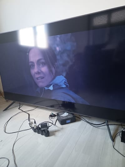 A large flat-screen television is mounted on a wall in a room located at 43.114° N, 1.618° E in the city of Pamiers, France. The television screen displays a scene from a movie featuring a woman with long brown hair, wearing a blue jacket, looking directly at the viewer. The television is connected to a black rectangular cable box, with a power strip and various cables leading to the cable box. The floor is made of light wood. The room is brightly lit, with a window visible in the reflection of the television screen.