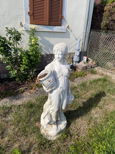 The image depicts a sunlit garden scene outside a residential building in Gerstheim, France. In the foreground, a white stone statue of a woman holding a basket with dried leaves stands on a stone base amidst overgrown grass. Behind the statue, a rose bush with yellow flowers is visible. To the right of the statue, a grey garden gnome and a smaller figure are placed near a chain-link fence. The building's exterior wall is textured and painted white, with a brown shuttered window. A drainpipe runs vertically along the wall. The ground is a mix of grass, gravel, and mulch. The overall impression is that of a well-maintained, albeit slightly untidy, outdoor space.