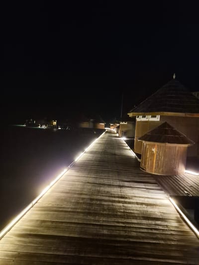 A photograph, rotated 180 degrees, depicts a night scene in Bodufolhudhoo, Maldives. A wooden structure, illuminated by a strip of light, runs along the right side of the image. To the left, dark water reflects the structure and other lights in the distance.