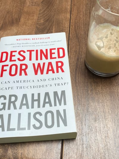 The scene is located in Ridgefield, United States, depicting a static arrangement on a medium brown wooden surface. A copy of the book "DESTINED FOR WAR" by Graham Allison, featuring a white cover with red and gray typography, is positioned on the left side of the frame. The title "DESTINED FOR WAR" is in red, with "NATIONAL BESTSELLER" and a quote attributed to "HENRY KISSINGER, former US secretary of state" visible above it. The author's name, "GRAHAM ALLISON," is in gray. To the right of the book, a clear glass contains a light brown, frothy beverage, approximately half full. The wooden surface exhibits distinct grain patterns and plank divisions. No individuals or active movements are present.