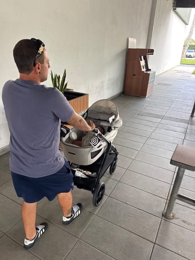 A man with short brown hair, wearing a grey t-shirt, dark blue shorts, and black and white sneakers, pushes a beige baby stroller. He is looking to his right. The stroller contains a baby. The man has tattoos on his legs. He is walking on a tiled pathway. Behind him, a wooden trash receptacle stands against a white wall. In the background, a car is visible on a street, and palm trees line the area. The scene appears to be in a modern, outdoor setting. It is daytime.