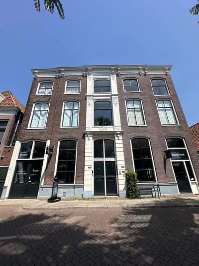 The image captures a section of a brick building on a sunny day in Zwolle, Netherlands. The building features several floors with numerous windows, some of which are adorned with white curtains, while others offer a clear view into the interior. Ornate white trim decorates the facade between the window levels, adding architectural detail. The ground floor appears to house businesses, with visible signage. One sign reads "MODULNOVA" and another appears to be for "VAN GALEN INTERIEUR" with Dutch text that translates to "FURNITURE AND INTERIOR". Below "VAN GALEN INTERIEUR" there is text that says "VAN GALEN". The building is situated on a cobblestone street, with shadows cast from an unseen source, indicating it is likely late morning or early afternoon. The sky is a clear, vibrant blue, suggesting pleasant weather. A small amount of greenery is visible, possibly a potted plant or bush near one of the windows. A street sign with the number "28" is visible on the facade. Another sign with the number "223" is visible on a window on the second floor. Another sign with the number "221" is visible on a window on the first floor.
