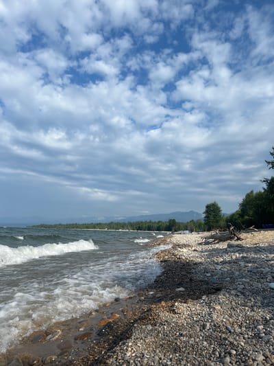 The image depicts a serene lakeside scene at Tolbazikha, Russia. The foreground is dominated by a rocky shoreline with waves gently lapping at the pebbles. The water is a deep blue-green, reflecting the overcast sky. In the distance, a dense forest lines the shore, meeting a range of misty mountains under a sky filled with scattered white clouds. A piece of driftwood rests on the beach to the right, with scattered tents and a small boat visible further down the shore. The overall impression is one of natural beauty and tranquility.
