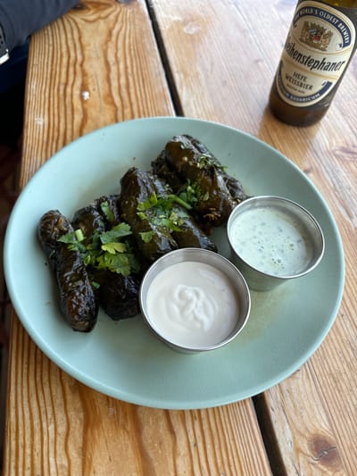 The image displays a dining setting on a light-colored wooden table with prominent grain, located in the Megilot Yam HaMelakh Regional Council, Palestinian Territory. Centered on a light green, round ceramic plate are several dark green, rolled items, consistent with stuffed grape leaves (dolma), garnished with chopped fresh green herbs. Two small, silver-colored metal ramekins are positioned on the plate; one contains a creamy, off-white sauce, and the other holds a pale green, liquid sauce with visible specks. In the upper right corner, a brown glass bottle of Weihenstephaner Hefe Weissbier is partially visible. The upper left corner shows a segment of a dark-colored sleeve, suggesting the presence of an individual at the table.