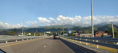 The image depicts a scenic view of a highway with a backdrop of majestic, snow-capped mountains under a clear blue sky dotted with fluffy white clouds. The sun appears to be setting or rising, casting long shadows and illuminating the scene with a warm, golden light. The highway, with its clear lane markings and guardrails, suggests a journey or commute.
On the highway, several cars are visible. In the foreground, a dark SUV is driving away from the camera. Further ahead, other vehicles can be seen, suggesting moderate traffic. The surrounding landscape is lush and green, with rolling hills leading up to the imposing mountain range.
On the right side of the highway, houses with red-tiled roofs and well-maintained yards are nestled amongst trees, indicating a populated area adjacent to the road. There are also streetlights lining the highway. A blue road sign with white arrows pointing upwards and to the sides is visible, indicating lane guidance. Another sign in the distance has Cyrillic text, which appears to read "РАЗВЯЗКА ЗАТУЛЕК" (Razvyazka Zatulek), likely indicating a road junction or interchange.
The overall mood is one of peaceful travel and the grandeur of nature. The clear weather and the impressive mountain vista create a sense of awe and tranquility. The presence of human infrastructure, such as the highway and houses, juxtaposed with the natural beauty of the mountains, highlights the harmonious coexistence of human settlement and the natural environment. The image captures a moment of serene travel, with the destination or journey being facilitated by the well-maintained road, leading towards or away from the captivating mountain landscape.