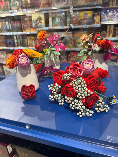 The image displays a retail store's display window or interior exhibit in Foligno, Italy, featuring multiple floral arrangements constructed entirely from LEGO bricks. The scene is set on a glossy, reflective royal blue surface, which occupies the lower and mid-ground.
Three white, vase-like objects are arranged in the mid-ground, each containing distinct LEGO flower bouquets. The leftmost vase holds a vibrant mix of orange, red, yellow (sunflower-like), and light purple LEGO flowers. A single red LEGO rose rests on the blue surface directly in front of this vase. The middle vase, positioned slightly behind and to the right, contains magenta-petaled flowers with light green stems and small light blue accents. The rightmost vase, also slightly elevated and further back, holds red, orange, and magenta LEGO flowers.
In the foreground, a large, horizontal bouquet of predominantly red LEGO roses is prominently displayed on the blue surface. This bouquet is interspersed with numerous clusters of small white LEGO pieces, simulating baby's breath. A single light pink LEGO rose is visible among the red roses, and a small blue and yellow daisy-like LEGO flower is located to the right side of this main bouquet. Reflections of the LEGO flowers and vases are visible on the blue surface.
The background, largely out of focus, reveals multiple shelves stocked with boxed merchandise, consistent with board games or similar products, behind a transparent barrier such as a glass display case or window. The lighting is artificial, suggesting an indoor retail environment. No human figures are present in the frame.