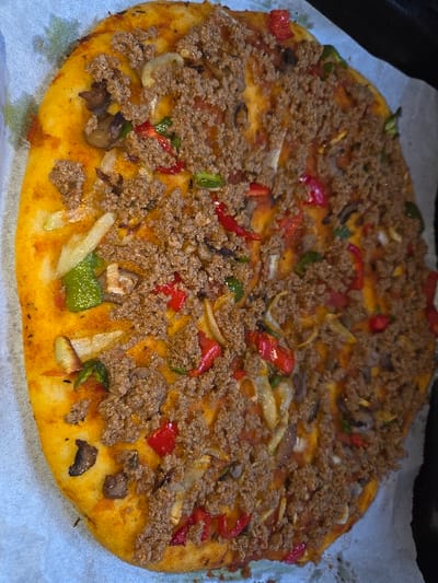 A circular, baked flatbread-style dish, consistent with a pizza or lahmacun, is depicted from an overhead perspective. The dish rests on a sheet of white parchment paper, which is partially visible within a dark baking tray. The base consists of a golden-brown crust. The surface is heavily covered with a uniform layer of finely crumbled, cooked ground meat. Scattered throughout the meat topping are visible pieces of diced red bell pepper, sliced green bell pepper, sliced white onion, and sliced dark brown mushrooms. A reddish sauce base is discernible beneath the toppings. The item appears recently cooked. No individuals or actions are depicted. The location is stated as Blainville-sur-Orne, France.