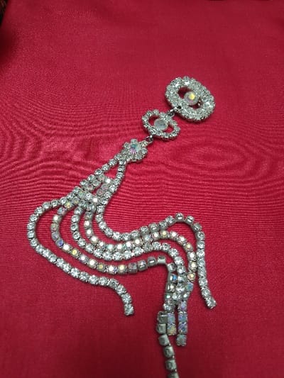 This image, captured in Horní Vltavice, Czechia, depicts a single piece of elaborate rhinestone jewelry positioned on a deep red, lustrous fabric background. The jewelry features silver-toned metal settings encasing numerous clear, round-cut rhinestones. Several larger, central stones throughout the piece exhibit an iridescent 'aurora borealis' coating, reflecting hues of pink, yellow, and blue.
The ornament is structured with three main segments. The uppermost segment is a large, elongated oval shape, bordered by small clear rhinestones, enclosing a prominent iridescent central stone. Directly below this is a smaller, interlocked teardrop or heart-shaped element, similarly outlined with clear rhinestones around its own iridescent center. These two top elements are connected by a short segment of clear rhinestones to a central anchor point. From this anchor point, approximately seven to eight flexible strands of clear rhinestones cascade downwards and towards the right of the frame. These strands are arranged in dynamic, curved lines, resembling a stylized tassel or flowing water. The individual rhinestones are uniform in size along each chain.
The background is a vibrant, solid deep red fabric, possibly satin or silk, characterized by a visible sheen and subtle folds, indicating it is a draped, soft material. The lighting is even and soft, emphasizing the sparkle of the rhinestones and the texture of the fabric. The perspective is a slightly angled overhead shot, focusing tightly on the jewelry. No people, other objects, or environmental context beyond the fabric are visible within the frame.