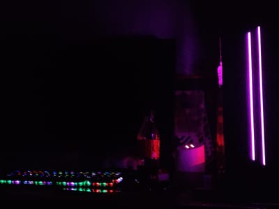 A dimly lit scene is captured in what appears to be a room, with a prominent focus on colorful LED lights. In the foreground, a keyboard with illuminated keys displays a spectrum of colors, including green, purple, blue, and red. To the right, three vertical neon pink lights stand out against the darkness. In the mid-ground, a bottle with a red label is visible, illuminated by a reddish-purple light. Behind the bottle, a dark structure with a ladder-like pattern is faintly illuminated, and to its right, a white object with two small pink lights is present. The overall atmosphere suggests a setting that is either a gaming room, a studio, or a space with decorative lighting. The image does not provide any context to determine its location in Roranwala, Pakistan.