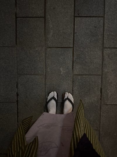 An overhead view captures the lower extremities of a person standing on a dark grey, rectangular tiled surface in the city of Yamanouchi, Japan. The individual wears white tabi socks, featuring a distinct split-toe, paired with dark-strapped traditional Japanese sandals. The lower portion of a garment, likely a kimono or yukata, is visible, consisting of a central light lavender fabric with a subtle embossed pattern. The outer panels of the garment display a striped pattern in olive-green and brown tones, with a dark, possibly purple, fabric section also present on the right side. The scene is illuminated by low ambient light.