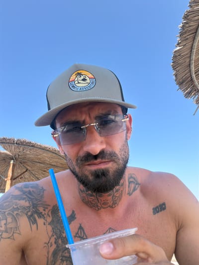 This media file is a close-up, low-angle shot of a person on a beach under a clear blue sky. The subject, a person with a beard and tattoos, is wearing a baseball cap and sunglasses. A straw from a drink is visible near their mouth, and they appear to be holding a translucent cup. The scene includes a portion of a straw umbrella overhead, suggesting a sunny outdoor setting. The person's tattoos are visible on their chest and shoulder. The baseball cap has a circular patch with the text "LIMNOS ADVENTURES" and an image of a palm tree within a sunset. Another tattoo on the person's upper chest appears to be text, possibly "BEOR". The time of day is likely midday, given the bright sky and the shadows. The overall mood suggests leisure and relaxation on a vacation.