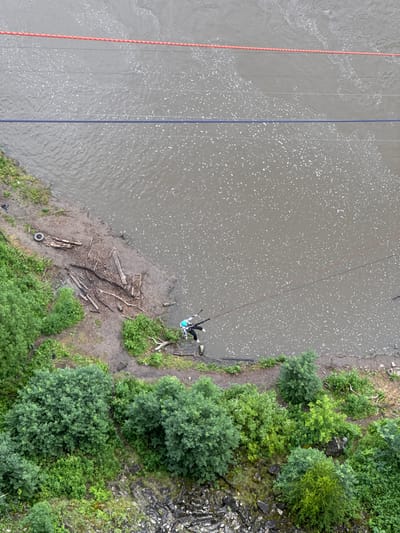 The image is an aerial shot of a person rappelling down a rope over a body of water. The person is wearing a teal and white outfit and black pants, with their legs extended and arms raised. They are attached to a rope that descends from above and appears to be suspended over a muddy bank littered with driftwood and a discarded tire. To the left of the water, there are two parallel ropes, one red and orange, and another dark blue. The water itself has a textured surface with numerous white specks. On the right side of the image, a lush green embankment with trees and rocks slopes upwards. The scene suggests an outdoor adventure activity, possibly taking place on a cloudy day given the diffused lighting and lack of harsh shadows. The location is identified as Kul'metovo, Russia.