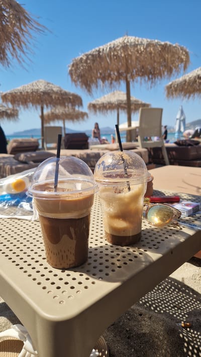 The image depicts a close-up of two iced coffee drinks with straws in clear plastic cups, placed on a beige plastic table. The drinks have a layered appearance, with a darker brown at the bottom and a lighter, frothy layer on top. In the background, several thatched umbrellas provide shade, and lounge chairs are visible. Beyond the beach setting, a body of water and a hazy shoreline are discernible under a clear blue sky.
The scene suggests a relaxed atmosphere, likely a sunny day at a beachside cafe or resort. The focus on the drinks and the presence of beach umbrellas and lounge chairs indicate leisure and vacation. The time of day appears to be daytime, given the bright sunlight casting distinct shadows from the table's holes onto the ground. The location is contextualized as Artemida Municipal Unit, Greece, which aligns with the typical imagery of Mediterranean beach resorts. There is no visible text in the image. The overall mood is one of tranquility and enjoyment.