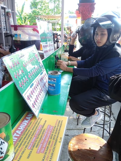 Two individuals wearing helmets sit at an outdoor stall in Kebondalem, Indonesia. The stall has a green countertop displaying menus and beverage options. One person is looking directly at the camera. The stall features an Arizona beverage dispenser and various canned goods. A sign details a price adjustment effective April 13, 2026, for "Es Teh Presiden" menu items, citing rising raw material costs. Both people appear to be sitting and waiting to order.
