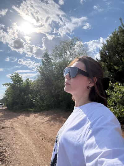 A person wearing sunglasses and a white t-shirt stands outdoors on a dirt path. Trees and blue sky with clouds form the background. The location is Doksha, Russia.