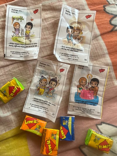 The image displays several wrappers from "Love is..." candies and two "Love is..." branded chewable candy bars scattered on a patterned fabric surface. Four of the wrappers are visible in their entirety, each featuring a cartoon illustration and text in Russian and English. The illustrations depict couples in various romantic scenarios, accompanied by phrases like "...having difficulty keeping your eye on the ball," "...sharing your whole future," "...someone to carry your skis," and "...seeing each other in a new light." The wrappers are dated 1997 and list "MINIKIM CARIBBEAN NV" as the copyright holder. Two "Love is..." candy bars, one predominantly yellow and the other orange, are also present. The scene does not contain any identifiable landmarks or elements that would suggest a specific location within the city of Новокатаево, Russia, or any other city. The fabric surface appears to be a blanket or bedspread with a light pink and orange striped pattern.