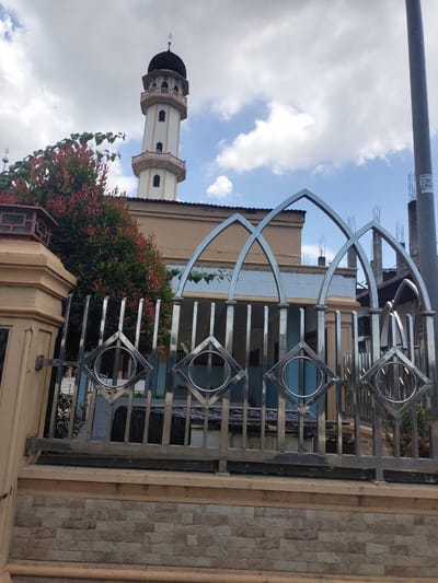 A mosque is visible through a stainless steel fence in Panteepanah, Indonesia. The fence has a low brick base, with metal bars and arched details. Behind the fence is a building, the mosque, with a tall minaret featuring a black dome. The sky is partly cloudy.