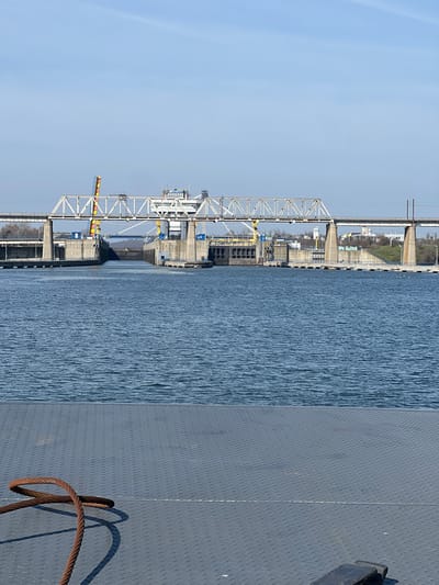 The image displays a panoramic view of an industrial waterway infrastructure located in Agigea, Romania, specifically a section of the Danube-Black Sea Canal. The perspective is from a low vantage point, likely a vessel's deck or a pier.

In the foreground, a dark grey, diamond-plate textured metal surface occupies the lower third of the frame. On this surface, in the lower left quadrant, a rusty, thick metal cable or rope is coiled, casting a distinct shadow.

The midground features a wide expanse of dark blue water, exhibiting numerous ripples across its surface.

The background is dominated by a large industrial complex spanning the waterway. This complex includes a massive concrete lock system with substantial vertical walls flanking a central channel through which water flows. A small, dark, triangular object is visible in the water within this channel. Above the lock and running parallel to it is a prominent steel truss bridge, supported by multiple robust concrete piers. This bridge carries a transportation route, likely a roadway or railway, across the canal. Industrial machinery, including yellow and red gantry cranes, are positioned within the lock complex. Directly behind the bridge structure on the right, a multi-story white building with numerous windows and a flat roof stands prominently, suggesting a control tower or administrative facility. On the far right, a vegetated landmass with additional structures is visible on a slight elevation. Several white gulls or similar birds are present on the water near the structures on the right side. The sky above is clear and light blue, devoid of clouds. The scene is illuminated by strong, direct sunlight, evidenced by the sharp shadows cast by objects. No human figures are discernible within the frame.