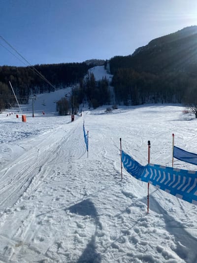 The image presents a sunlit ski slope within the ski resort area of La Salle-les-Alpes, France. The foreground features a snow-covered surface marked with numerous ski tracks and indentations. To the right, a blue and white striped safety banner, exhibiting a chevron pattern, is stretched between red poles. A separate blue mesh netting on black poles is situated further left in the mid-foreground. Shadows are cast upon the snow, indicating bright sunlight.
In the midground, the ski slope ascends, flanked by scattered trees and bushes. Multiple orange protective pads are visible on poles or obstacles along the left edge of the slope. To the left, a ski lift system is operational, showing several towers with wires extending upwards and empty chairlifts suspended. Numerous small, distant figures, identified as skiers, are visible descending various sections of the slope.
The background consists of steep, forested mountains. Dense coniferous forests cover substantial portions of the mountain slopes, with cleared ski runs discernible higher up. The sky is clear blue, and intense sunlight emanates from the upper right, producing visible lens flare. The overall scene depicts an active alpine ski resort on a clear day.