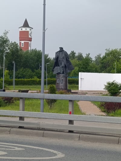 The image captures a statue of a man in a trench coat and hat, standing on a pedestal, in the city of Krasnaya Pakhra, Russia. Behind the statue, a tall, red brick tower with a conical roof and a clock face is visible, along with a white wall and lush green trees. A metal guardrail and a road with markings are in the foreground. The sky is overcast, suggesting a cloudy day.