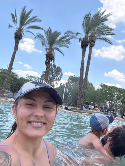 The image shows a woman in a body of water. She is wearing a cap and swimsuit. A man and child are visible near her. The location appears to be in Emek HaMaayanot Regional Council, Israel, indicated by the presence of palm trees, typical of that region. People are visible in the background, suggesting a pool or hot spring environment.