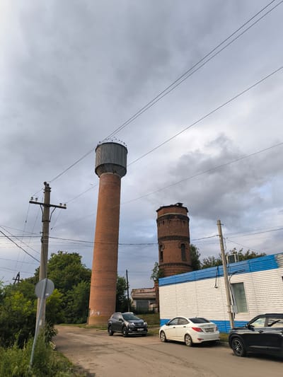 The image depicts an outdoor scene in Uzunovo, Russia, featuring two distinct water towers. The taller water tower, made of red brick, is prominently positioned in the left half of the frame. It has a cylindrical shape with a metallic, cone-shaped roof and is surrounded by an array of electrical wires and a utility pole. A smaller, older water tower, also constructed from red brick, stands to the right, appearing somewhat dilapidated with visible damage to its upper section.
In the foreground, a paved road runs horizontally across the image. Several vehicles are parked along the road. On the left side of the road, a dark gray SUV is parked, and a white sedan is parked to its right. Further to the right, a black SUV is parked. A white and blue building, possibly a temporary structure or a small industrial building, is situated behind the parked cars. Lush green trees and vegetation are visible in the background, framing the water towers and the surrounding area. The sky is overcast with heavy clouds. The scene appears to be a semi-industrial or utilitarian area within the town.