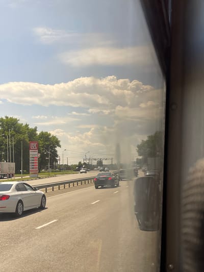 The image depicts a view from inside a vehicle, likely on a highway, with the camera oriented vertically. The scene is a roadside with a clear sky filled with large, white cumulus clouds, suggesting a sunny day. The road is asphalt with visible white lane markings, and it appears to be a multi-lane highway with several cars driving in the same direction. Trees line the side of the road, and in the distance, there are more cars and what appear to be streetlights and possibly buildings.

A prominent object in the scene is a gas station sign for "ЛУКОЙЛ" (LUKOIL), a Russian multinational energy corporation. The sign displays fuel prices, with various types of fuel listed. Next to the LUKOIL sign, there are more structures associated with a gas station, including pumps.

The activity taking place is vehicular traffic on a highway. The presence of the gas station indicates it's a location where drivers might stop for fuel. The time of day appears to be daytime, given the bright sunlight and clear sky, likely late morning or afternoon. The overall impression is a typical scene of travel on a highway in a suburban or semi-rural area of Russia. There are no people clearly visible in the image. The mood is one of everyday travel.