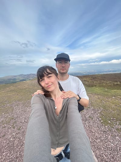A low-angle, selfie-style photograph captures a couple standing on a rocky, grassy hillside with a panoramic view of undulating, green hills and a distant town under a partly cloudy sky. A woman is in the foreground, smiling and looking at the camera, with a man behind her with his arm around her shoulder. The woman is wearing a gray, button-up cardigan and denim shorts, while the man is wearing a white t-shirt, glasses, and a black baseball cap. The setting appears to be a rural or mountainous area, possibly within or near the city of Penicuik, United Kingdom, given the resemblance to the Scottish landscape. The lighting is soft, suggesting an overcast day.