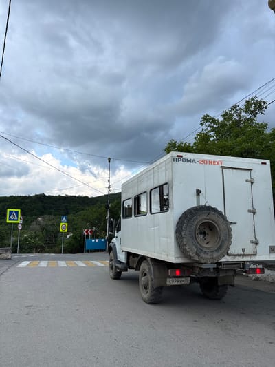 The image shows a white truck with a spare tire mounted on the back, parked on the side of a road. The license plate is K979YM82. "ПРОМА-20 NEXT" is written on the side of the truck. A pedestrian crosswalk is visible, with a corresponding sign. Additional signs indicating a speed limit of 40 are nearby, along with the direction of a turn. A forested hillside rises in the background, partially obscured by electrical wires. The sky is overcast. There is no evidence to suggest this image is in Zaprudnoe, Ukraine.