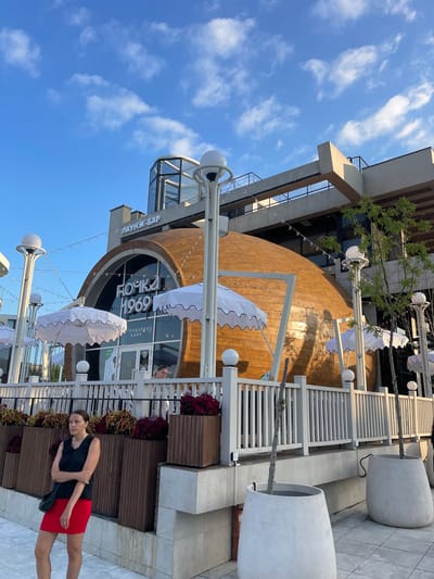 A woman in a black sleeveless top and a red skirt stands on a paved area next to a building with a distinctive curved, wooden facade. She is wearing a black crossbody bag. Behind her, there is a white railing, planters filled with plants, and large, white planters. In the background, the building features a large, circular window with text visible. Above the building, a blue sky with scattered white clouds is visible. The lighting suggests it is daytime, possibly late afternoon, with the sun casting some shadows. The overall scene appears to be an outdoor seating area for a cafe or restaurant. Text visible on the building includes "ПАУНЖ-БАР", "БОЧКА 1969", and "ЛЕГЕНДАРНОЕ КАФЕ". The location is Ivanovskaya, Russia.