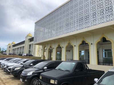 In Reuleut Timu, Indonesia, a modern mosque with distinctive golden domes and an intricately patterned facade is depicted under an overcast sky. The mosque features arched entrances and a pale yellow exterior. In the foreground, a row of parked vehicles, primarily SUVs and pickup trucks, are visible in a paved parking lot. The vehicles are a mix of black and white. The scene is set against a backdrop of the mosque and a grassy slope.