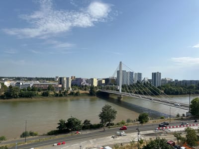This is an eye-level, medium shot photograph taken on a sunny day, showing a wide, muddy river with a modern suspension bridge crossing it. The bridge features a tall, white pylon with cables extending to the bridge deck, which is carrying vehicular traffic. On the far bank of the river, a dense urban landscape is visible, comprising a mix of apartment buildings of various heights and architectural styles, some featuring colorful facades. Trees and greenery are interspersed between the buildings and along the riverbank. In the foreground, a two-lane road runs parallel to the river, with several cars driving on it, including a red car and a black SUV. A sidewalk runs between the road and the river, with a few scattered trees and streetlights. There is also some construction work visible in the foreground with barriers and materials. A lone figure can be seen walking on the sidewalk near the bridge. The sky is clear blue with scattered white clouds. The scene depicts an urban environment with a prominent river and a modern bridge, likely within the city of Nantes, France, given the architectural style and the presence of the Pont de l'Île de Nantes, which this bridge resembles.