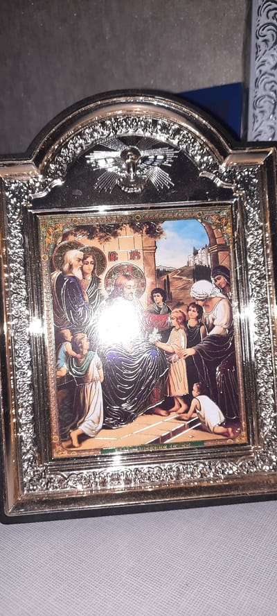 A framed religious icon depicts a scene of Jesus with children. Jesus, centrally located and emanating a bright light, is seated and surrounded by several individuals. To his left, two figures with halos are present: one older man with a white beard and a blue robe, and a woman with a dark veil. To Jesus' right, a group of children are gathered. One girl in a light-colored dress offers a flower to Jesus, while a young boy kneels before him. Another child in a blue garment is positioned to Jesus' left, leaning forward. The background shows an arched architectural element, suggesting an outdoor setting with structures and a hilly landscape under a blue sky. The frame is ornate and silver-toned, with decorative motifs. There is no specific indication of the location being Bocani, Moldova within the image itself.
