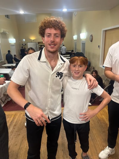 The image depicts two males, an adult and a child, standing together and smiling at the camera in an indoor community hall in Bergenfield, United States. The adult male, positioned on the left, has reddish-brown curly hair and wears a white short-sleeved collared shirt with black trim and a black "NY" embroidery on the left chest, black pants, a silver chain, and a black digital wristband on his left arm. His left arm is around the child's shoulders. The child, positioned on the right, has reddish hair and wears a black yarmulke, a white short-sleeved t-shirt with "SONNETI LONDON" text, black joggers, and grey sneakers. His right hand is placed on his hip.
The background shows a blurred gathering of multiple individuals, some seated at white-covered tables. The room features light beige walls, a light-colored wooden floor, and a white ceiling equipped with rectangular fluorescent and wall-mounted lighting fixtures. A wall-mounted air conditioning unit is visible on the left wall, and a circular clock hangs on the right wall. A green beverage can is visible on a table in the lower-left corner.