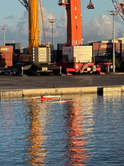 The image depicts a detailed view of the Papeete harbor in Papeete, France, likely during morning or late afternoon due, to the warm, directional lighting. In the foreground, a body of water shows ripples and distinct reflections of industrial equipment. Two individuals are present in slender boats, resembling racing kayaks or canoes. One person, wearing a red top, is in a red boat positioned closer to a concrete quay. Another individual, wearing a lighter-colored top, is in a white boat slightly further away. A third boat, white with red accents, is partially visible.

The midground features a light gray concrete quay extending horizontally across the frame. The number "6" is prominently displayed on the vertical face of the quay.

The background is dominated by an active port environment. Two large mobile harbor cranes are central. The crane on the left is primarily yellow and gray, featuring "LIEBHERR" branding on its counterweights, and appears to be a large Liebherr mobile crane. The crane on the right is predominantly orange and red, with "TEREX GOTTWALD" and "sat nu" visible on its base, indicating a Terex Gottwald crane. A red spreader or lifting attachment hangs from its boom. Numerous shipping containers, stacked in various configurations, fill the area behind and between the cranes. Visible container markings include "MARFRET" and "CMA CGM". Several tall light poles with multiple fixtures are also present across the port yard. The sky above is partially blue with scattered white clouds.