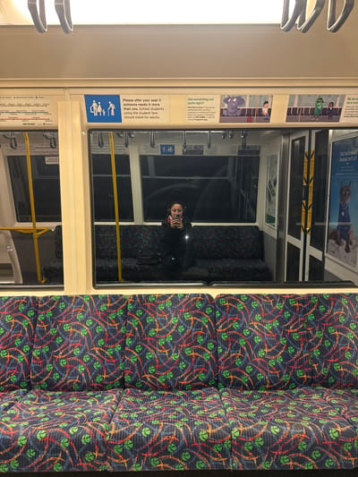 The image depicts the interior of a train carriage in Bassendean, Australia. The carriage has patterned navy blue seats with orange, red, blue, and green designs. A person is seated and is using their phone. Posters and signage are visible on the walls, including information about fare concessions and seat etiquette. The METRONET logo is displayed, indicating the train network.