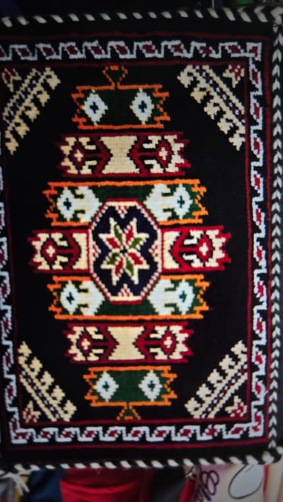 This is a close-up, high-angle shot of a woven rug with a black background and intricate geometric patterns in red, white, yellow, green, and orange. The rug appears to be handmade, possibly a tribal or kilim rug, given the style of the patterns which are symmetrical and repeat across the surface. The central design features a star-like motif within an octagonal border, surrounded by elongated, vertical decorative elements that resemble stylized figures or amulets. The edges of the rug have a decorative border with smaller repeating patterns. The lighting is somewhat dim, and the image has a slight blur, suggesting it was taken with a handheld camera in low light conditions or at a distance. There are no people or animals visible, and the focus is entirely on the rug itself. The setting is not clearly defined, but the texture and style of the rug might suggest a traditional or ethnographic context. There is some out-of-focus background visible on the right side, including what appears to be fabric in red and possibly a part of a sign with partial text that is illegible. There are no discernible time of day or weather cues. The overall impression is one of craftsmanship and cultural artistry.
