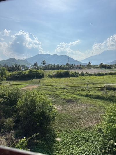 The image shows a panoramic view of a rural landscape in Van Hung, Vietnam, under a bright, partly cloudy sky. In the foreground, lush green vegetation dominates, with a variety of trees and bushes. A dirt path is visible in the middle ground, bordered by grassy fields and a few power lines. In the distance, a cluster of houses and palm trees are nestled amongst rolling hills, with a larger mountain range forming the backdrop. A single bird is seen soaring in the sky. The scene depicts a serene, natural environment, characteristic of the Vietnamese countryside.