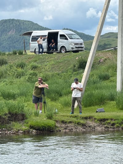 In Kazanovka, Russia, a group of individuals are engaged in filming activities near a body of water. In the foreground, a man wearing sunglasses, a green t-shirt, and shorts stands next to a tripod with a camera mounted on it. To his right, another man in a white t-shirt and a black baseball cap holds a drone controller, appearing to operate a drone. In the background, a silver van with its rear door open is parked on a grassy slope. Two men are visible inside and near the van. The environment is characterized by lush green grass, rolling hills in the distance, and a cloudy sky. Power poles are present on the right side of the frame. The scene suggests a filmmaking or photography expedition in a natural, rural setting.