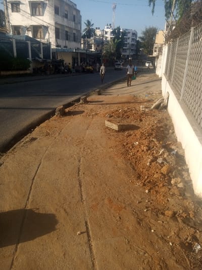 The image depicts an urban street scene in the city of Ganjoni/Shimanzi ward, Kenya, under clear daytime conditions. In the foreground, a heavily degraded dirt and gravel sidewalk runs parallel to the road on the right. This sidewalk features uneven terrain, visible depressions or tracks, and scattered piles of reddish-brown earth mixed with concrete fragments and debris. Several broken concrete curb sections are present on the sidewalk's edge. A long, dark shadow extends diagonally across the lower left portion of the sidewalk.
Separating the sidewalk from the main thoroughfare is a partially intact curb line. The midground shows a paved, dark asphalt road. Two adult males are walking away from the camera on the road, towards the background. The male on the left wears a light-colored shirt and dark trousers, while the male on the right wears a white t-shirt and dark trousers, carrying a green bag in his left hand.
On the left side of the road, a row of multi-story buildings, predominantly light-colored, line the street. Ground-level facades reveal various shops or stalls, some with awnings, where several individuals are visible standing or seated. A dark metal fence or gate fronts one of the buildings on the immediate left. Further down the road, multiple parked vehicles are visible.
The background consists of additional urban architecture, including taller residential or commercial buildings. Several green palm trees are interspersed among the structures. A tall, reddish-brown cellular tower is prominent on the left side of the distant skyline. The sky above is clear and light blue.