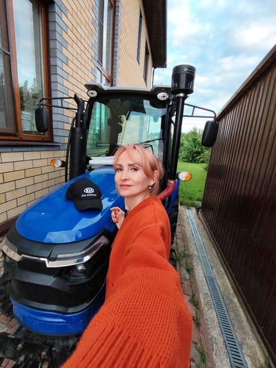 A woman with blonde hair, wearing an orange sweater, takes a selfie with a blue tractor. A black baseball cap with a white Kia Motors logo is placed on the hood of the tractor. The tractor is parked next to a building with a brick facade and a corrugated metal fence. The scene is outdoors, with green grass and trees visible in the background. The sky is partly cloudy. This scene is located in Kstovo, Russia.