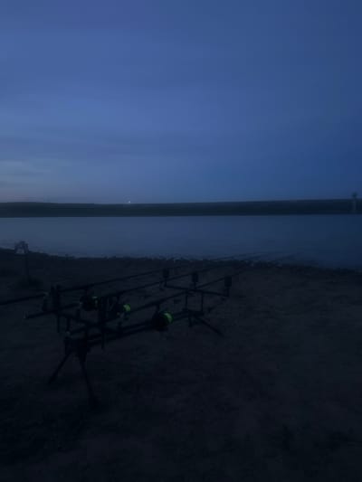 The photograph depicts a waterside scene, possibly at dusk or dawn, with a dark blue sky transitioning into a dimly lit body of water. Three fishing rods are set up on a stand along the shore, their lines extending into the water. The shore is dark and appears to be composed of dirt or sand. In the distance across the water, a faint light is visible. The location is likely near Trastikovo, Bulgaria, given the prompt, although specific identifiable landmarks are not apparent in this image.