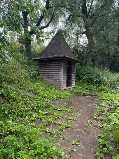 In Nikulskoye, Russia, a small, weathered wooden structure with a pyramidal roof stands amidst lush greenery. The building, constructed from horizontal wooden planks, features a single entrance with an ornate metal grate. The surrounding area is a dense forest with a path made of wooden planks that appears damp and overgrown with grass and small plants. The trees are tall and their branches, laden with leaves, create a canopy overhead, suggesting a secluded, natural setting. The scene is peaceful and appears to be after a rainfall, as indicated by the wetness of the ground and foliage. There are no people or animals visible in the image.