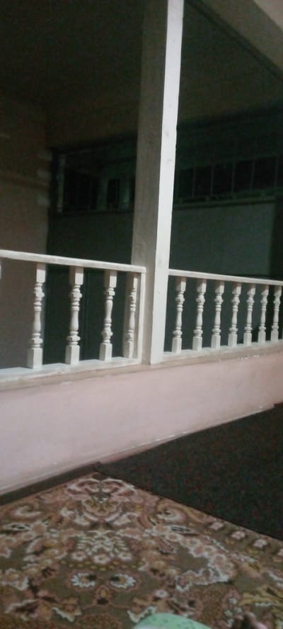 The image depicts an indoor scene, likely a residential area, in Marhamat, Uzbekistan. The foreground is dominated by a patterned carpet with earthy tones and intricate floral designs. Behind the carpet, a light-colored wall extends upwards. A wooden railing with white spindles and a light-colored top rail runs horizontally across the midground, suggesting a balcony or a raised platform. Beyond the railing, the environment appears to be an interior hallway or room with a large, cream-colored column positioned centrally. The background is dimly lit, revealing what seems to be a dark wall or entryway with multiple windows or openings. The overall impression is one of a domestic interior, possibly an apartment or house, with traditional decorative elements such as the carpet and railing.