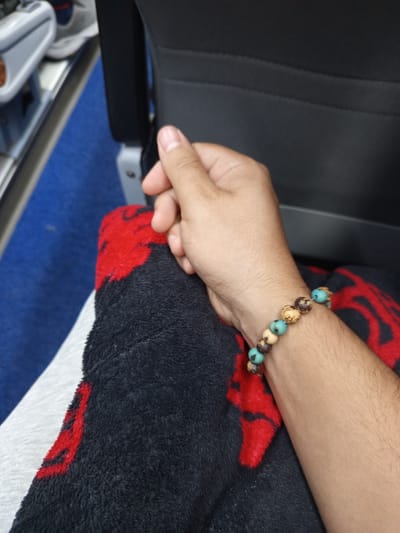 The image shows a close-up of a person's arm and hand, resting on a black and red patterned blanket. The person is wearing a beaded bracelet on their wrist. The background suggests an indoor setting, possibly public transportation, with a blue carpet visible. There is no discernible information within the image to indicate a location in Bucaramanga, Colombia.